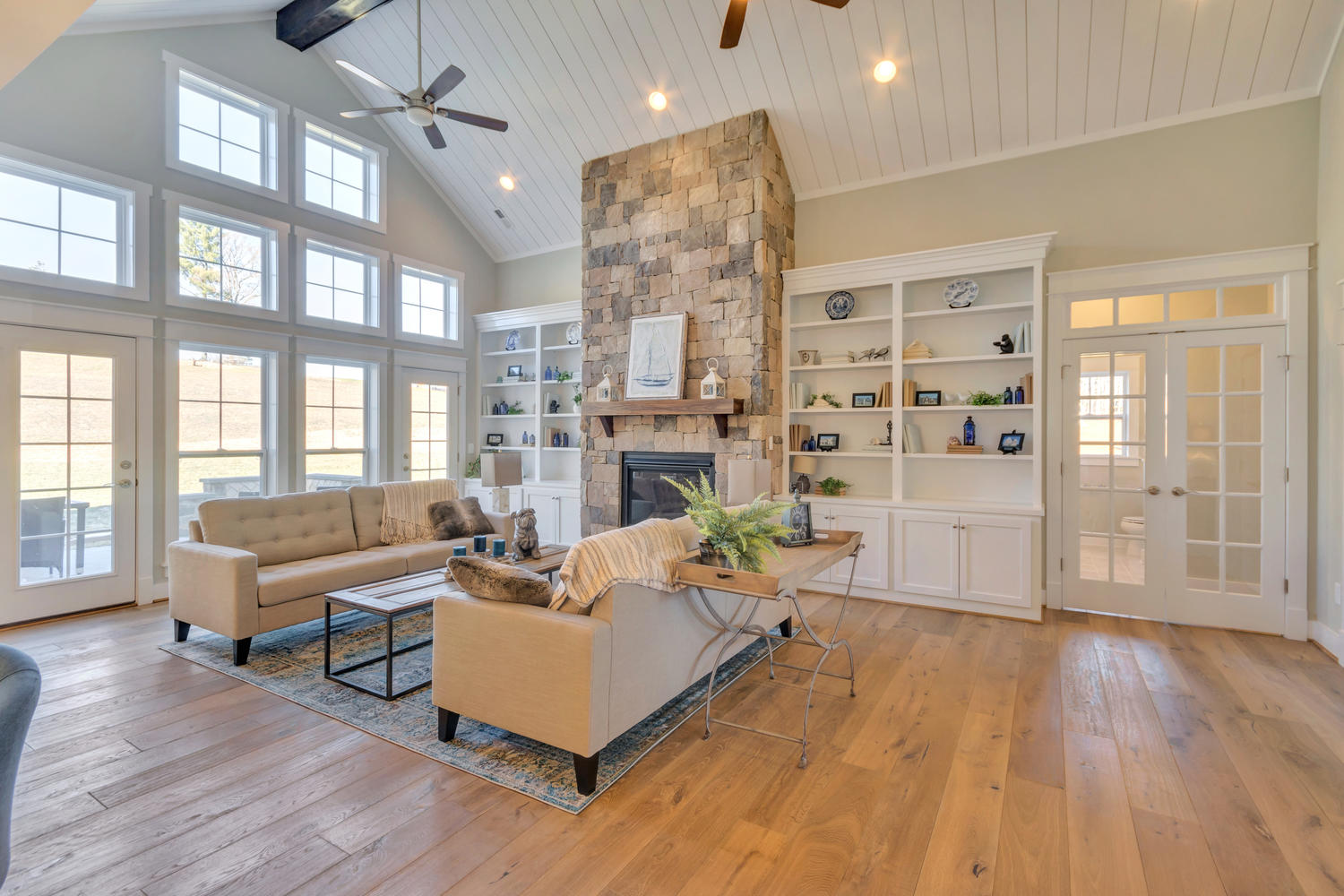 Craig Builders great room in Oak Hill Farm. A large bank of windows overlooks a small patio. Couches are arranged around a central stone fireplace.